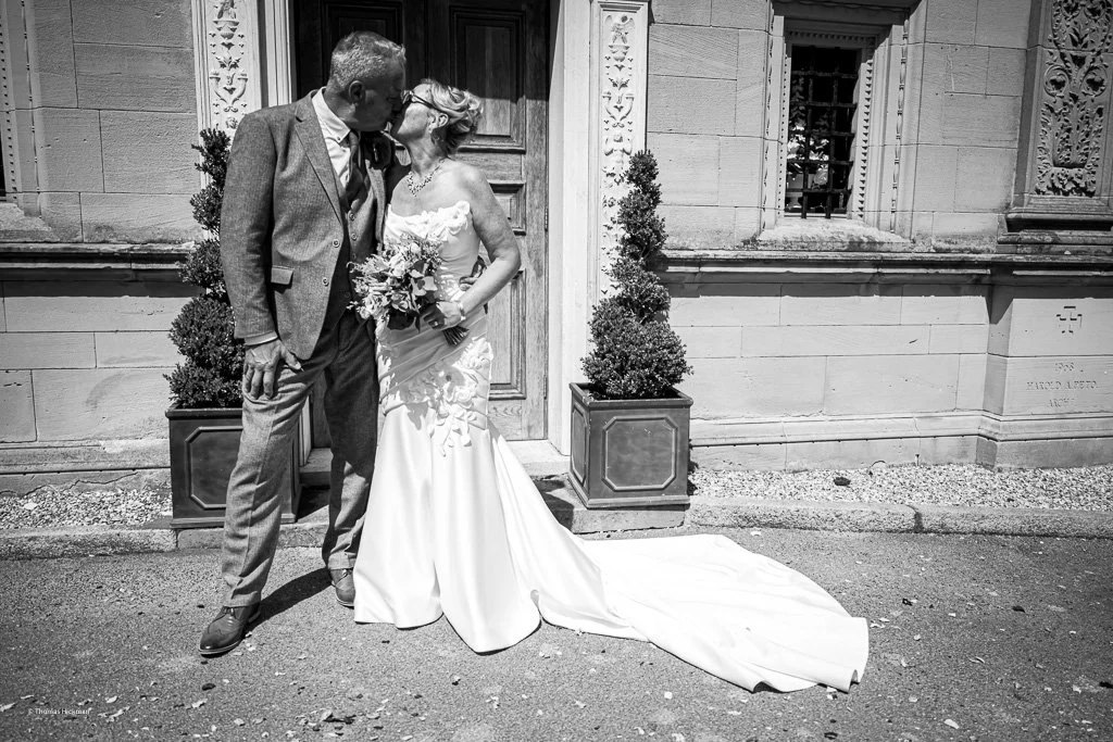Black and white photo of a bride and groom sharing a kiss outside a building with decorative architectural details. The bride is holding a bouquet and wearing a long wedding gown, while the groom is in a suit. Two potted plants flank the door behind 