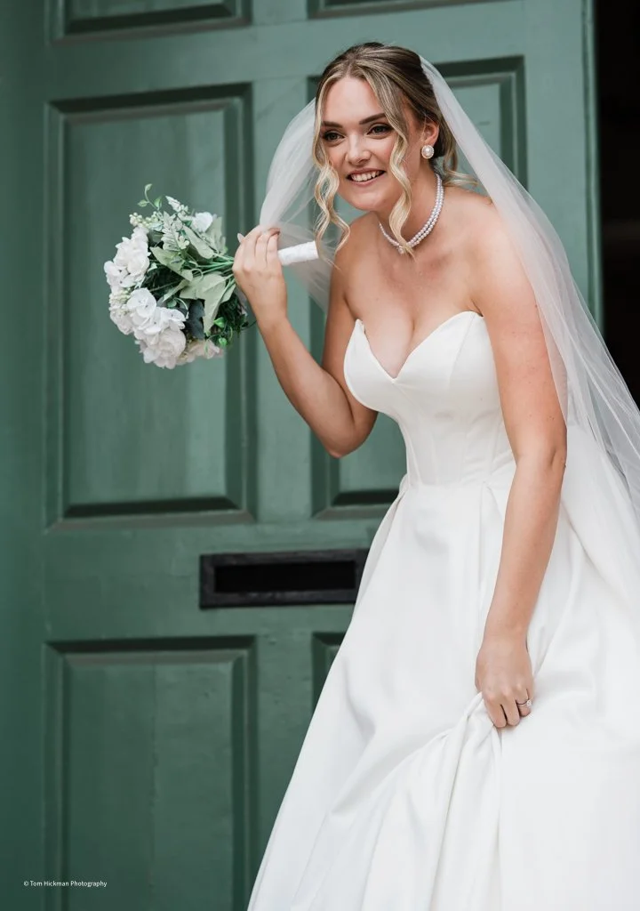 Bride in a white wedding dress holding a bouquet of flowers, smiling in front of a green door.