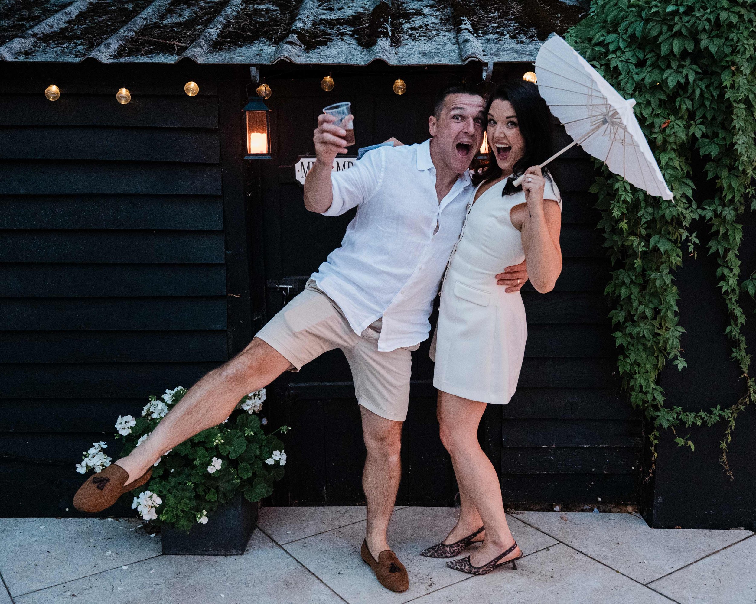 A man and woman dressed in white clothing standing outside against a black wooden wall with a small roof, string lights, and greenery. The man is holding a drink and jumping, while the woman is holding a white umbrella and smiling. They are standing on a stone sidewalk, with white flowers at their feet.