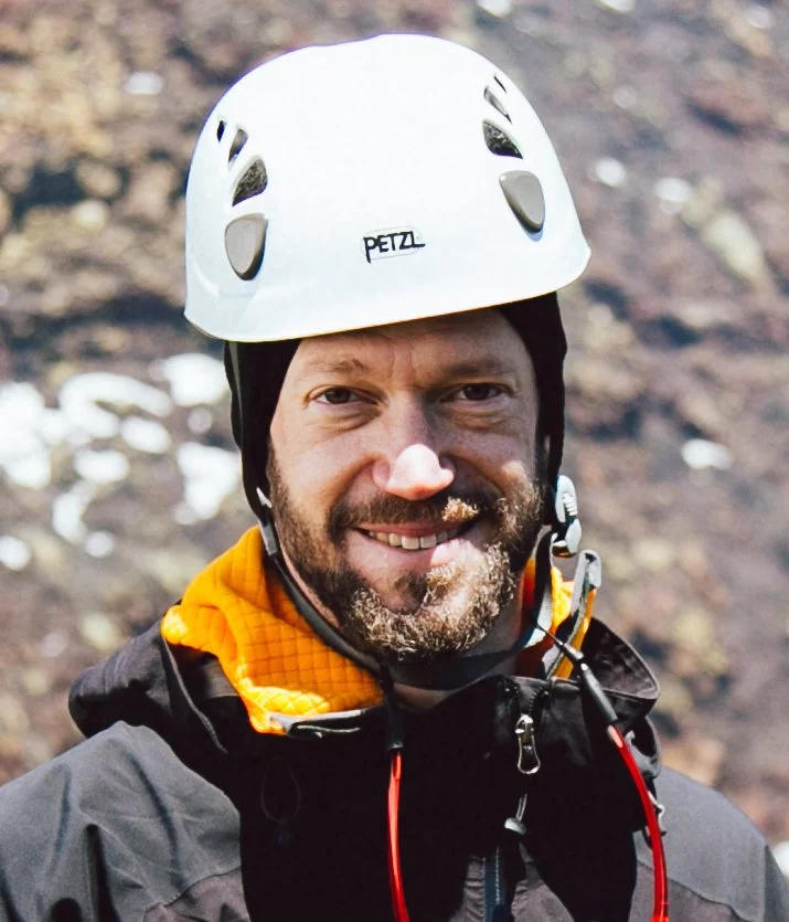 Man smiling wearing a white Petzl climbing helmet and outdoor gear, outdoors in a rocky environment.