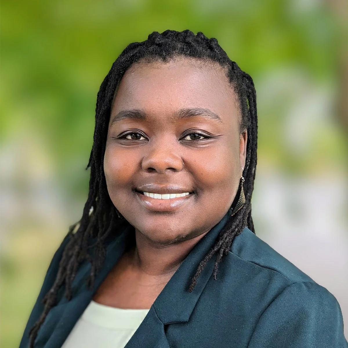 Close-up portrait of a smiling woman with dreadlocks, wearing a dark blazer and earrings, outdoors with blurred green background.