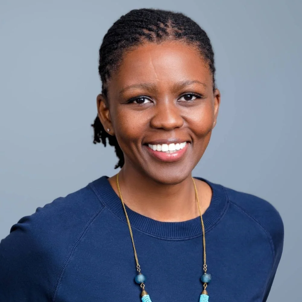 A smiling woman with natural short dreadlocks, wearing a blue shirt and a long beaded necklace, against a plain gray background.