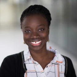 Portrait of a smiling young woman with dark skin, curly hair pulled back, wearing a black jacket over a white shirt with orange and black stripes, in an indoor setting.