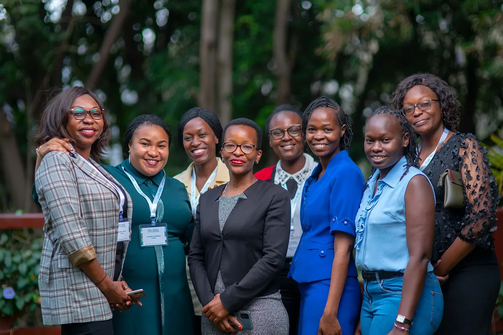 Group of nine women standing outdoors, smiling, in professional attire, with trees in the background.