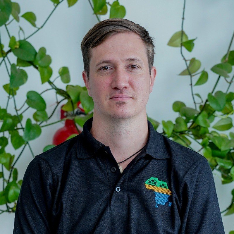 A young man with brown hair, wearing a black collared shirt with a colorful logo, standing in front of green leafy plants and a white wall.