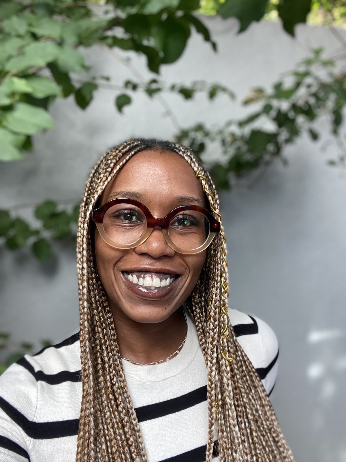 A woman with long blonde braids, wearing glasses and a striped shirt, smiling in front of a green leafy background.