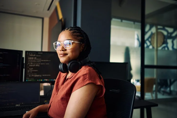 A woman with glasses and headphones sits at a desk in front of monitors in an office.