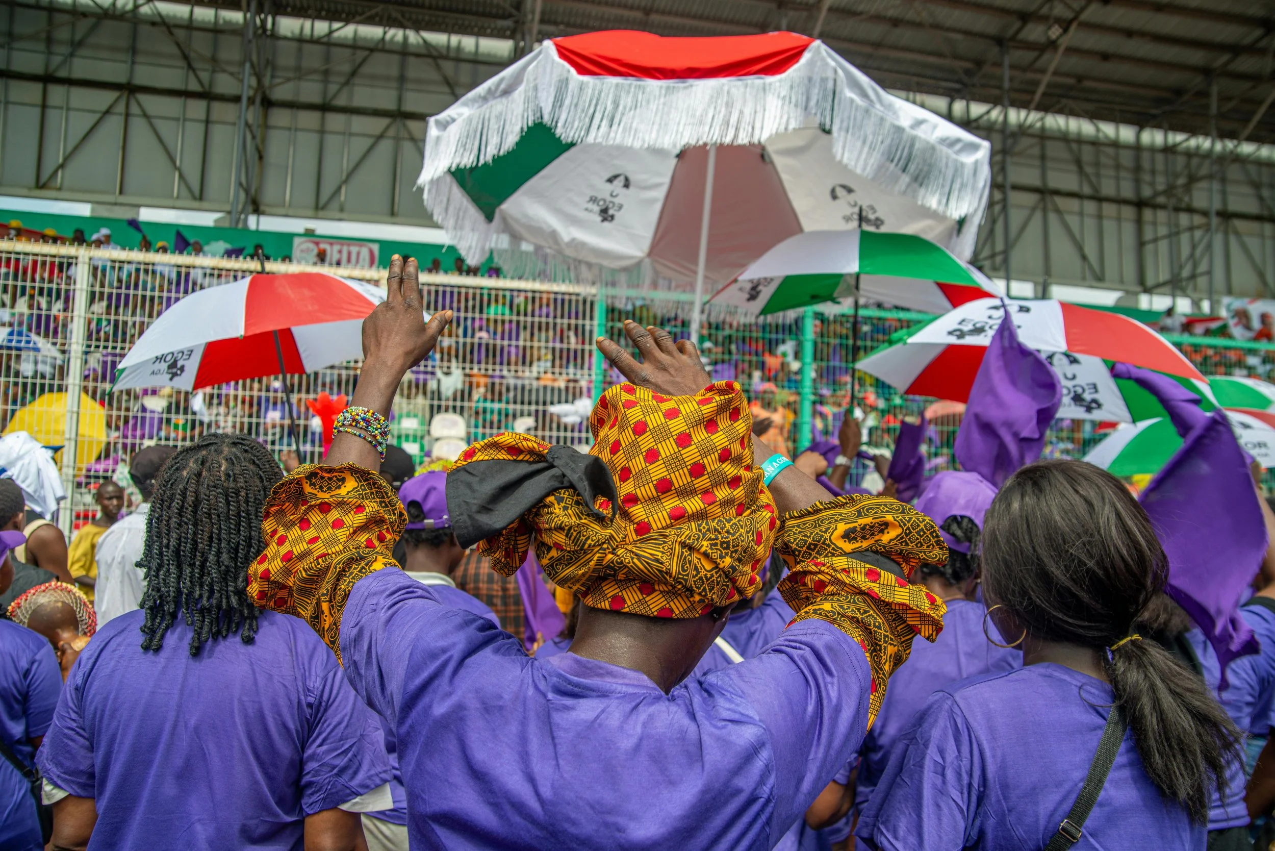 A crowd at a stadium during an event, with many people wearing purple shirts, holding umbrellas in red, white, green, and purple, and some people wearing traditional headscarves.