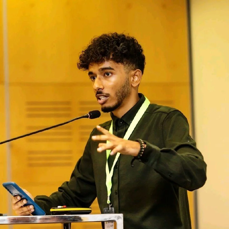Young man with curly hair and a beard speaking at a conference, holding a phone in his left hand, wearing a black shirt, gesturing with his right hand, standing at a podium with a microphone.