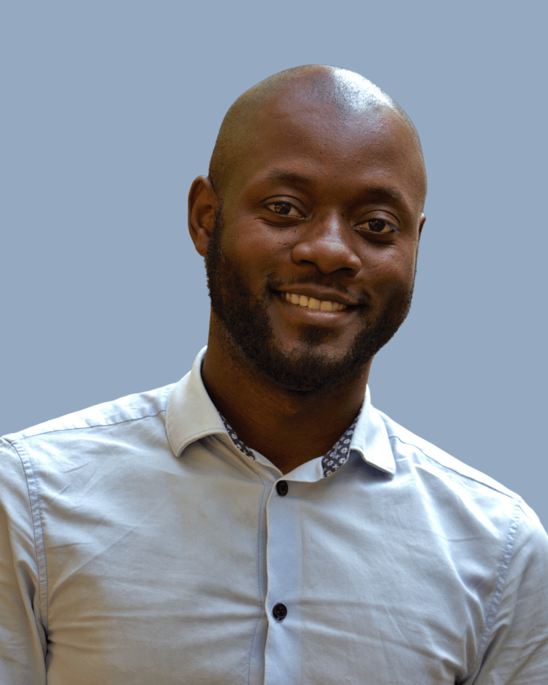 A smiling man with a bald head and a beard, wearing a light-colored dress shirt with buttons, standing against a plain light blue background.