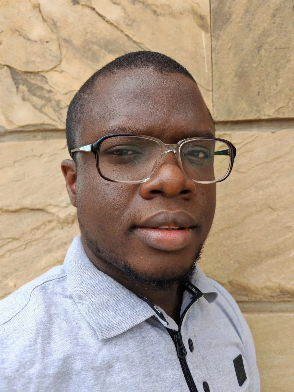 A young man with glasses and a short haircut taking a selfie against a stone wall background.