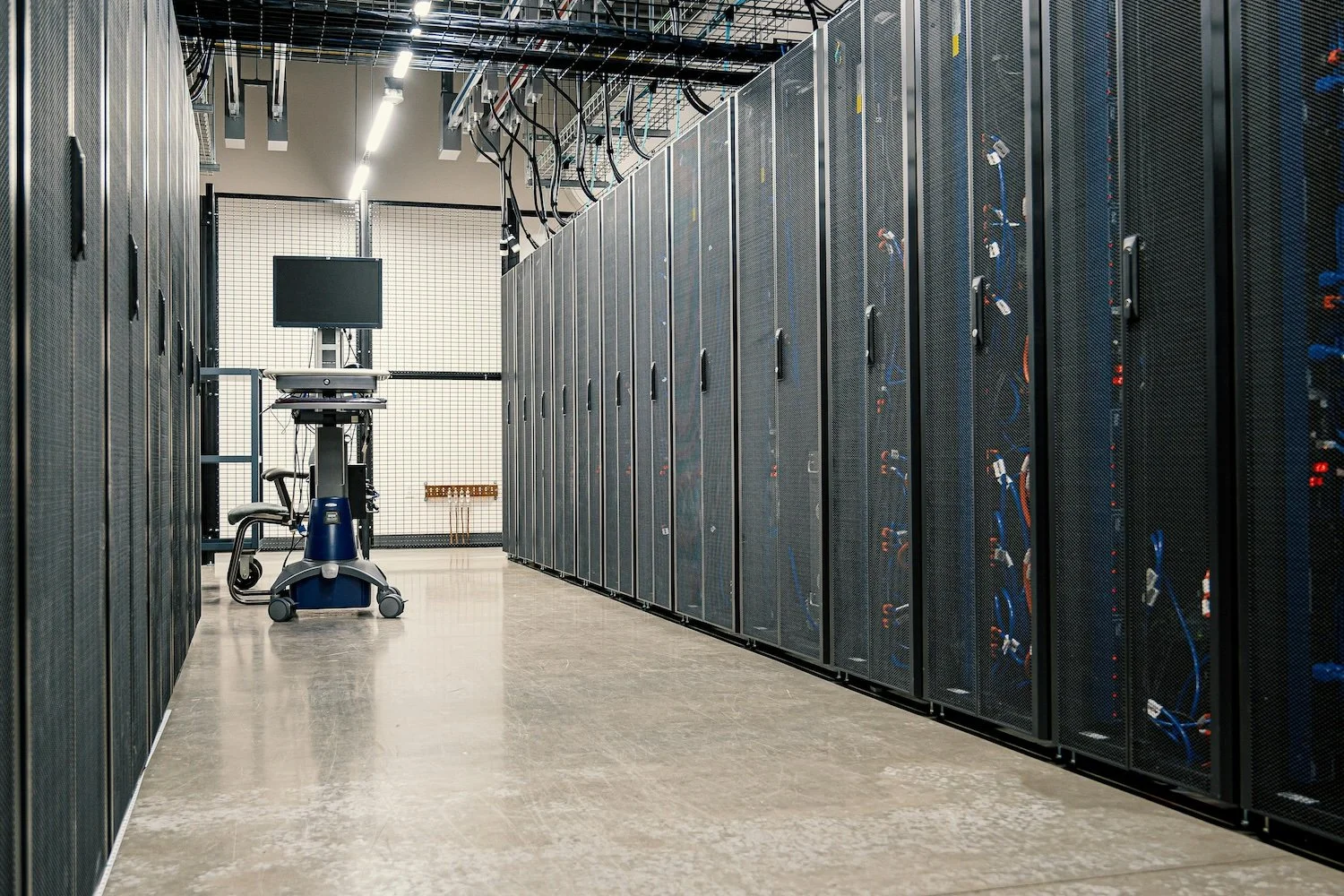 A server room with rows of black server racks on both sides, a mobile work station with a computer monitor, and a wire management panel at the back.