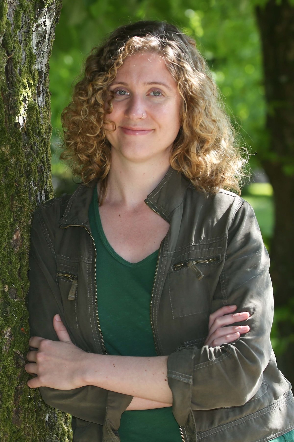A woman with curly blonde hair and a nose piercing stands outdoors next to a moss-covered tree in a green park, smiling and crossing her arms.