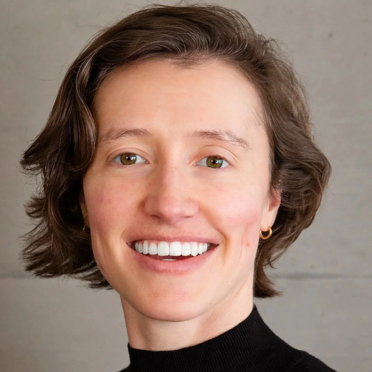 Close-up portrait of a smiling woman with light skin, short wavy brown hair, green eyes, and wearing a black turtleneck and small gold hoop earrings.