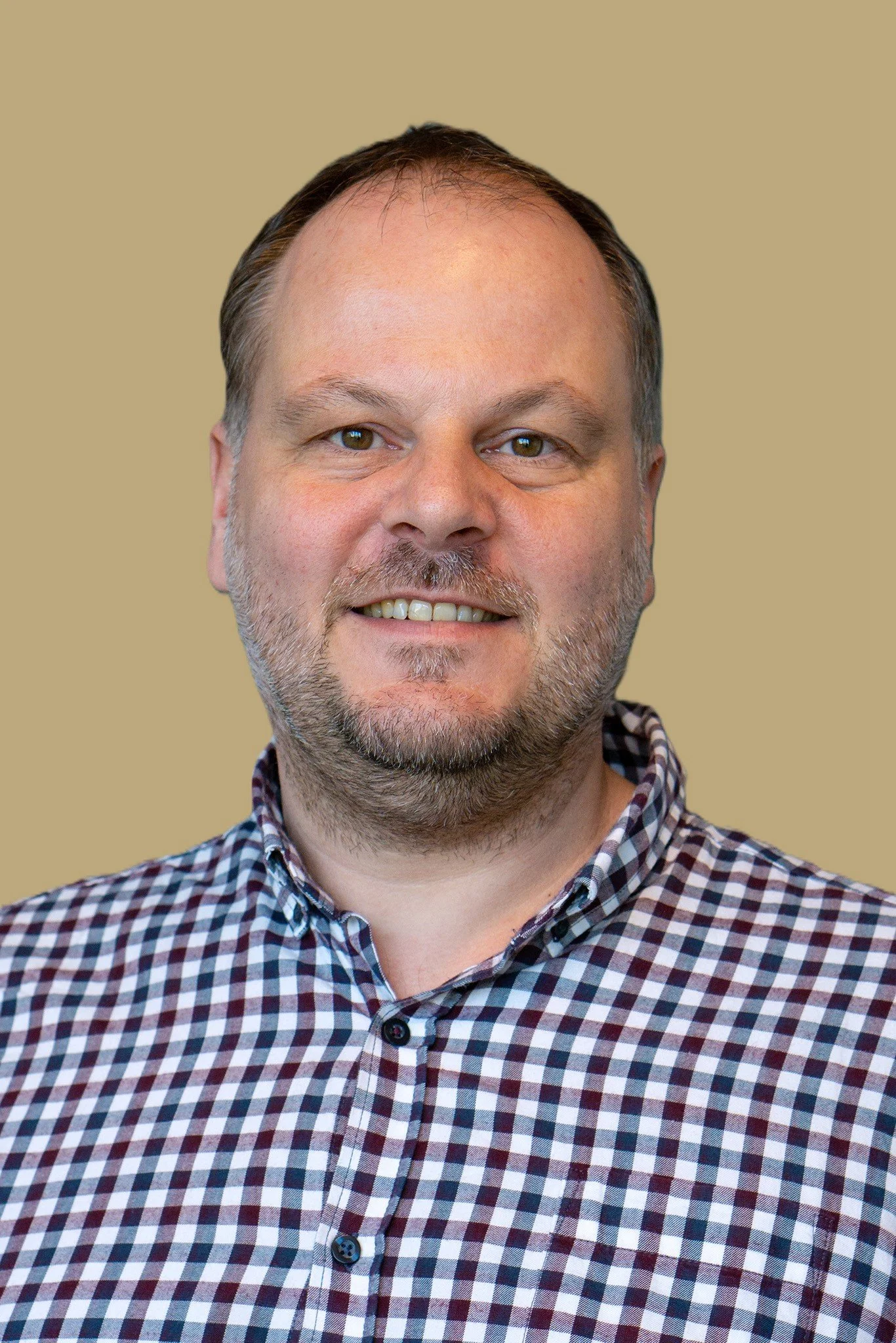 Portret van een man met kort haar, baard en een glimlach, dragend een geruite blouse, tegen een beige achtergrond.