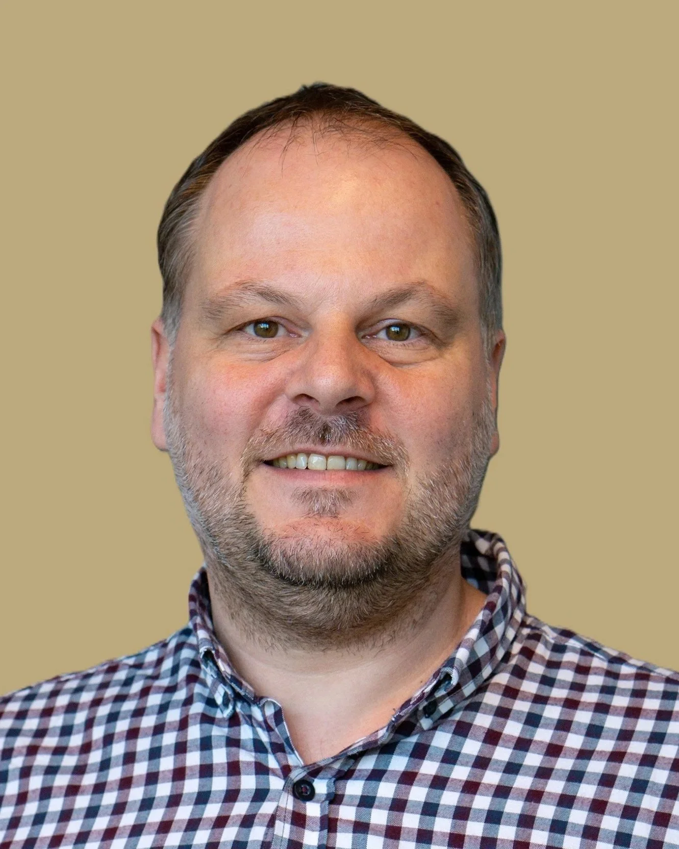 Portret van een man met een glimlach, korte haar, een baard en een geruite shirt, tegen een effen beige achtergrond.