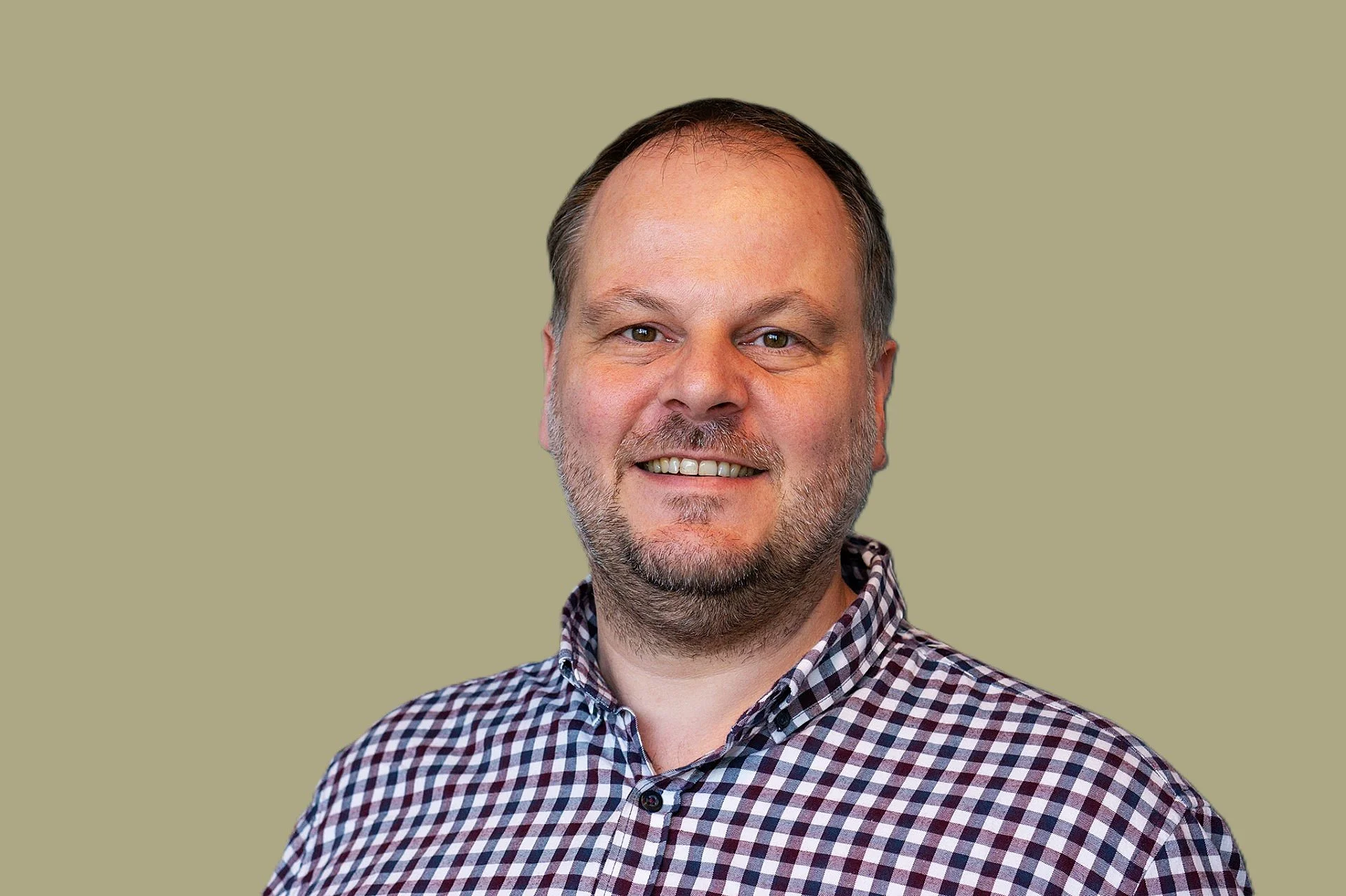Portret van een lachende man met een lichte huid, korte baard en haar, gekleed in een ruitjesoverhemd, tegen een effen groene achtergrond.