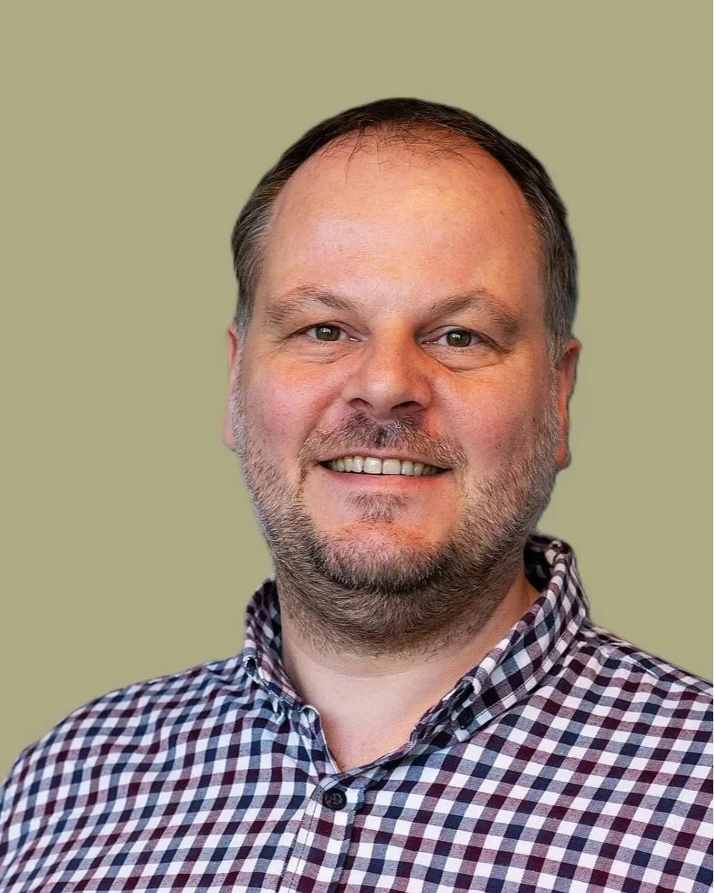 Portret van een man met een glimlach, korte baard en gestreept overhemd, tegen een effen lichtgroen achtergrond.