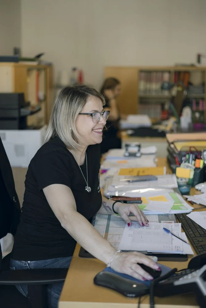 Donna che lavora sorridendo in un ufficio con scrivanie e scaffali pieni di documenti e materiali.