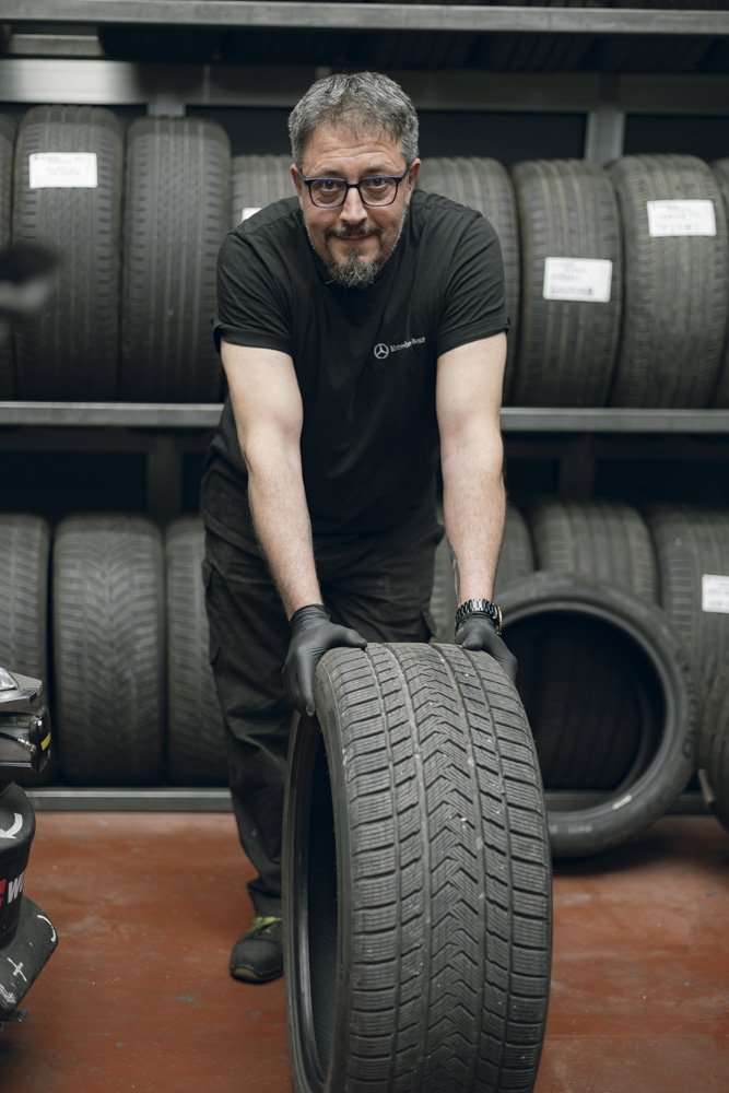 Uomo con occhiali e barba che tiene un pneumatico in negozio di pneumatici, sfondo di scaffali con più pneumatici.