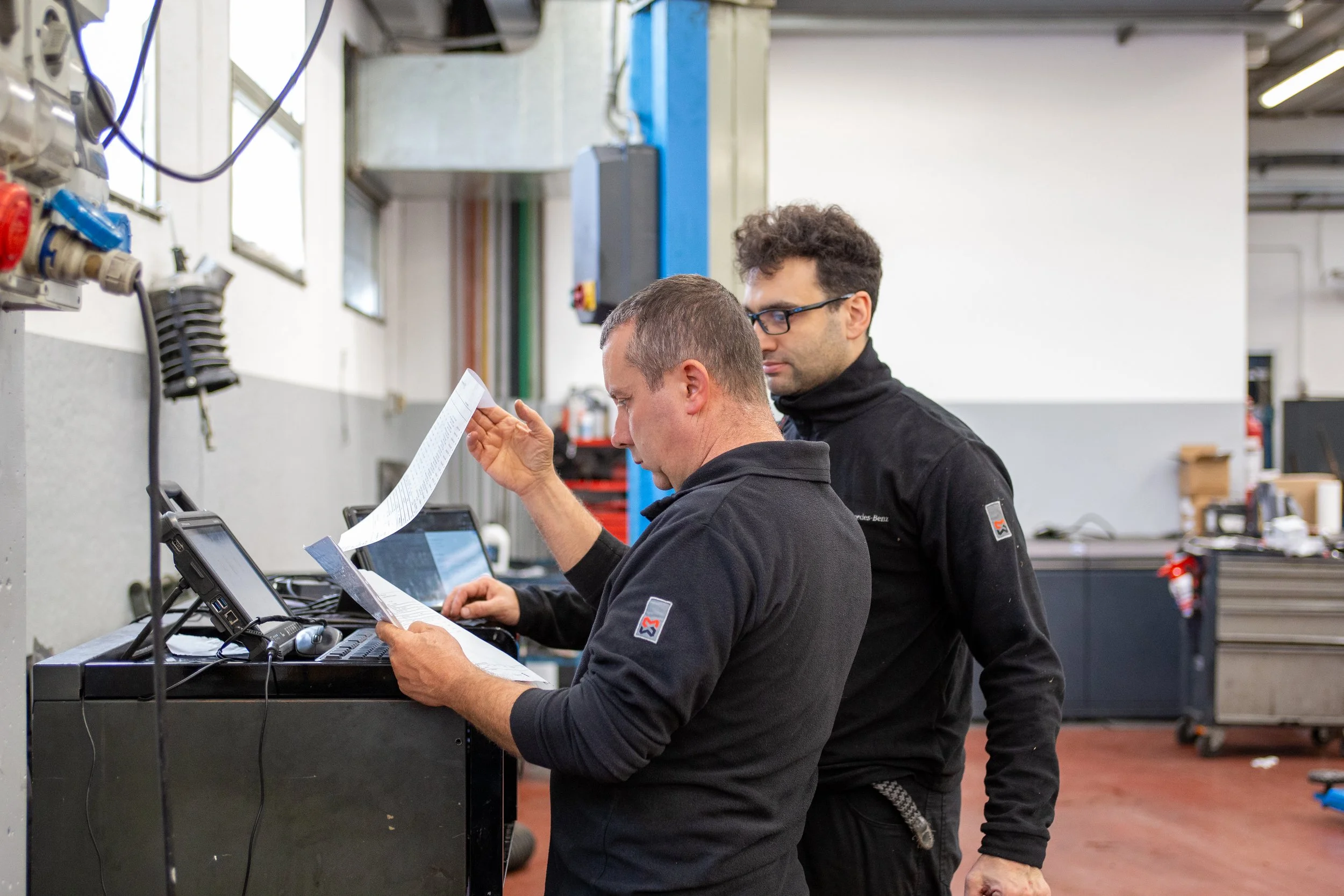 Due uomini in abbigliamento da lavoro discutono e guardano documenti e dispositivi elettronici in un'officina o garage.