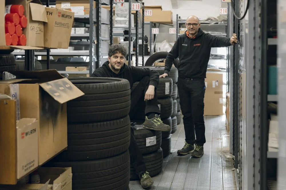 Due uomini in un magazzino tra scaffali e pneumatici, uno seduto e uno in piedi, entrambi sorridenti