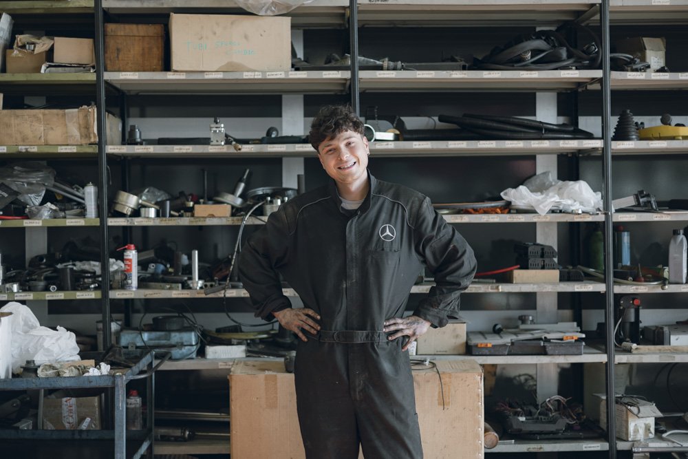 Meccanico sorridente in officina con scaffali di attrezzi e parti meccaniche sullo sfondo.