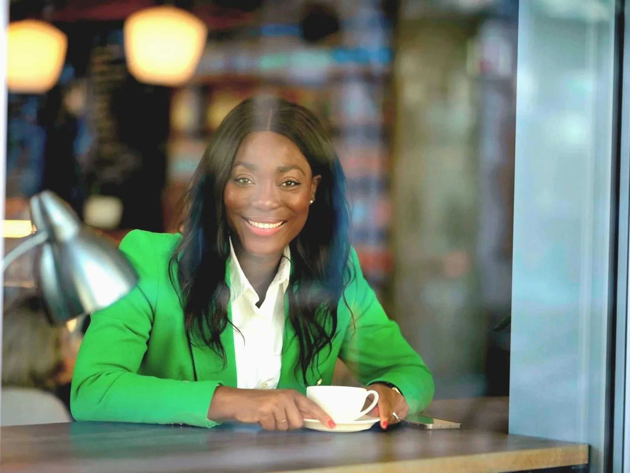 A woman with dark hair and dark skin, smiling, wearing a green blazer and white shirt, sitting at a table in a cafe, holding a white coffee cup.