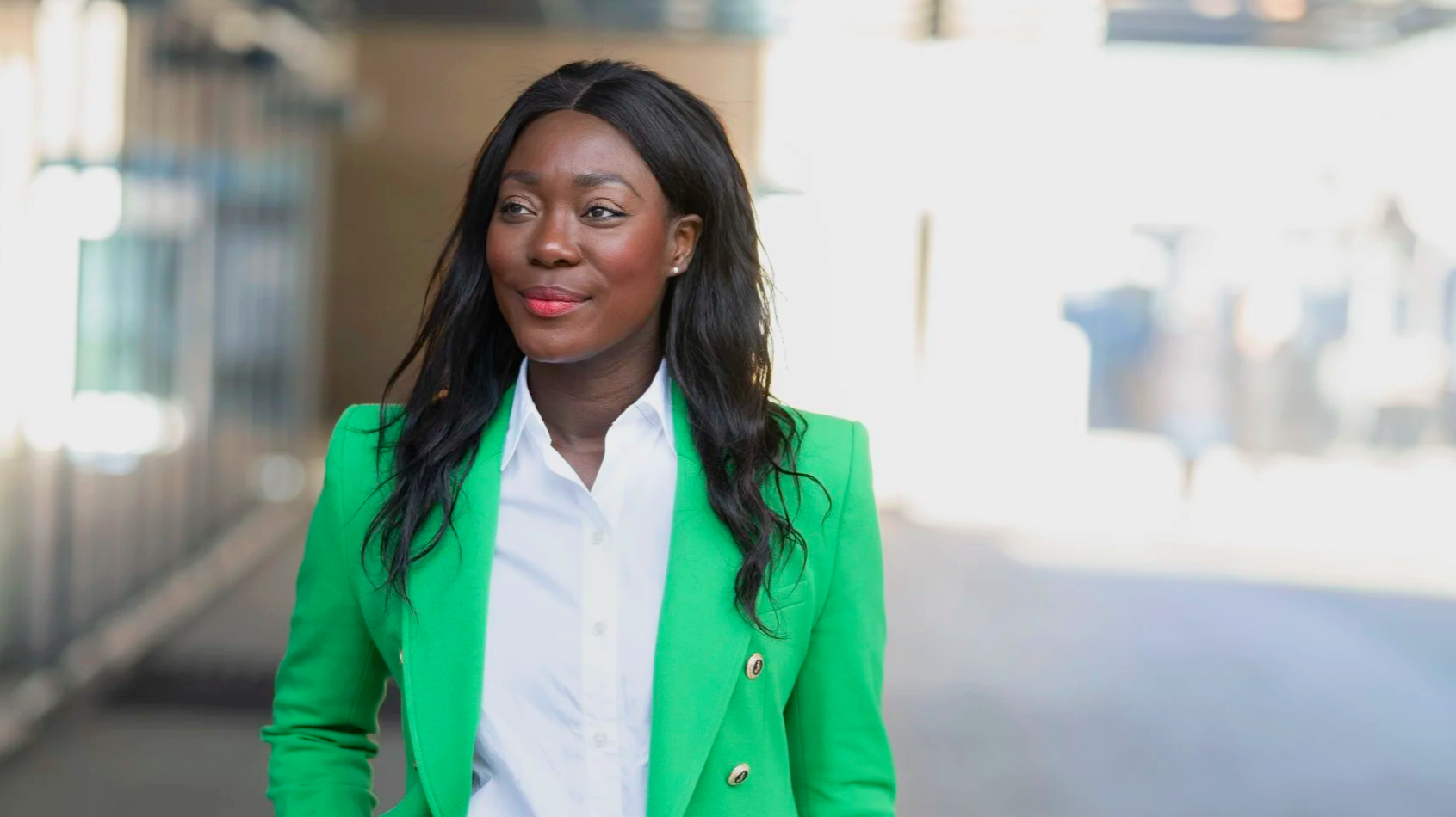 A woman with long black hair wearing a bright green blazer and white shirt stands outdoors, smiling softly. The background is blurred, with bright sunlight and buildings.
