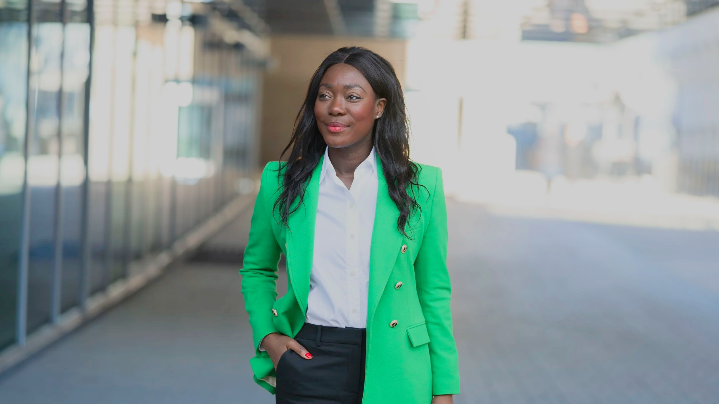 A woman with dark hair wearing a bright green blazer and black pants, standing outdoors with a modern building in the background.