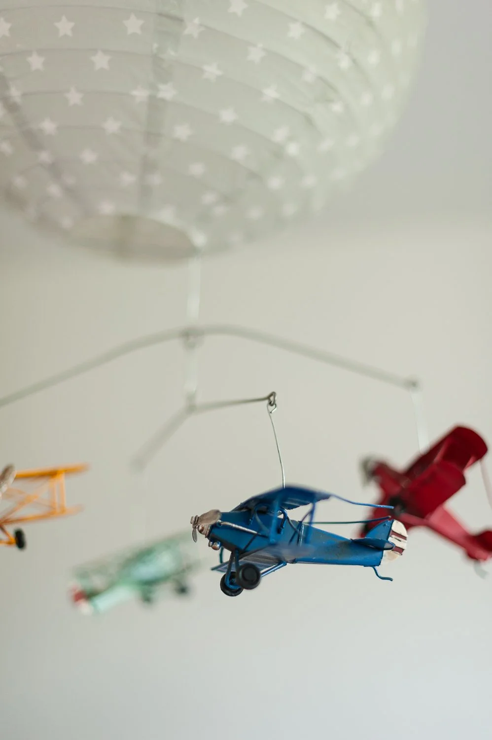 Miniaturni modeli letal, ki visijo z vrvic, vključno z izrazitim modrim modelom letala v ospredju, na beli luči, belem lampijonu s pikicami, mobile za otroke