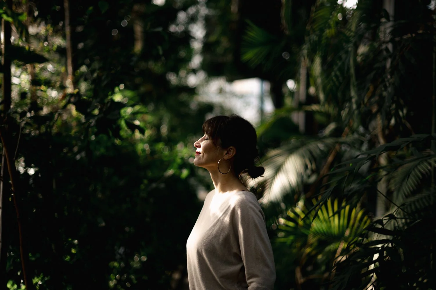 Ženska obsijana s soncem v svetlem puloverju z zaprtimi očmi v botaničnem vrtu. 