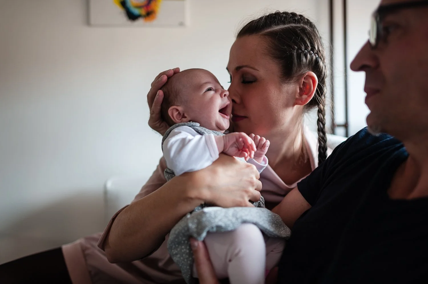 Ženska in moški držita nasmejano deklico, pri čemer ženska poljublja otrokov nos, blizu okna v prijetnem domačem okolju. ženska s francosko kito, novopečena mami.
