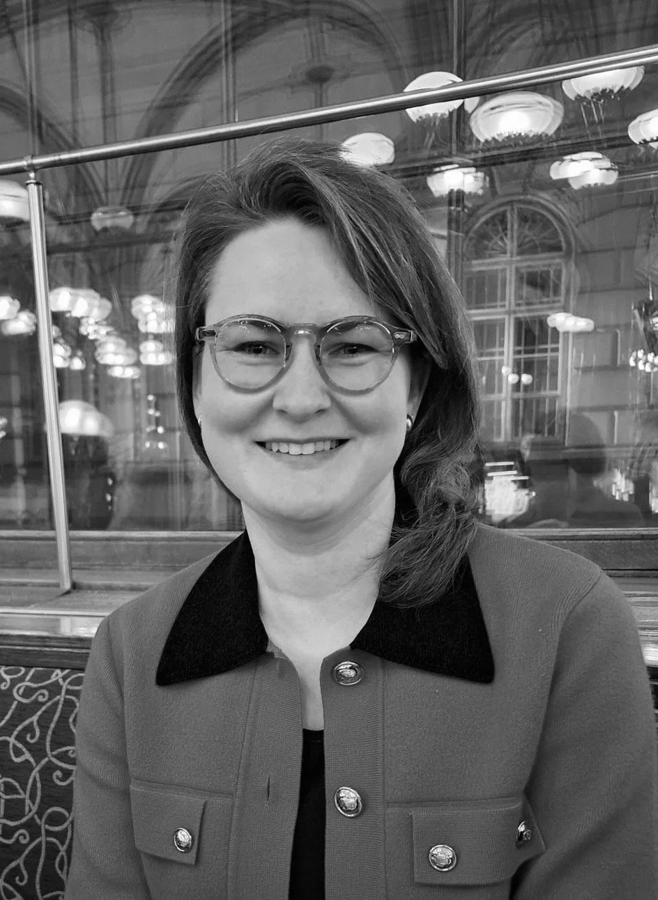 A woman with shoulder-length hair, wearing glasses and a jacket, smiling indoors with decorative lighting and large windows in the background.