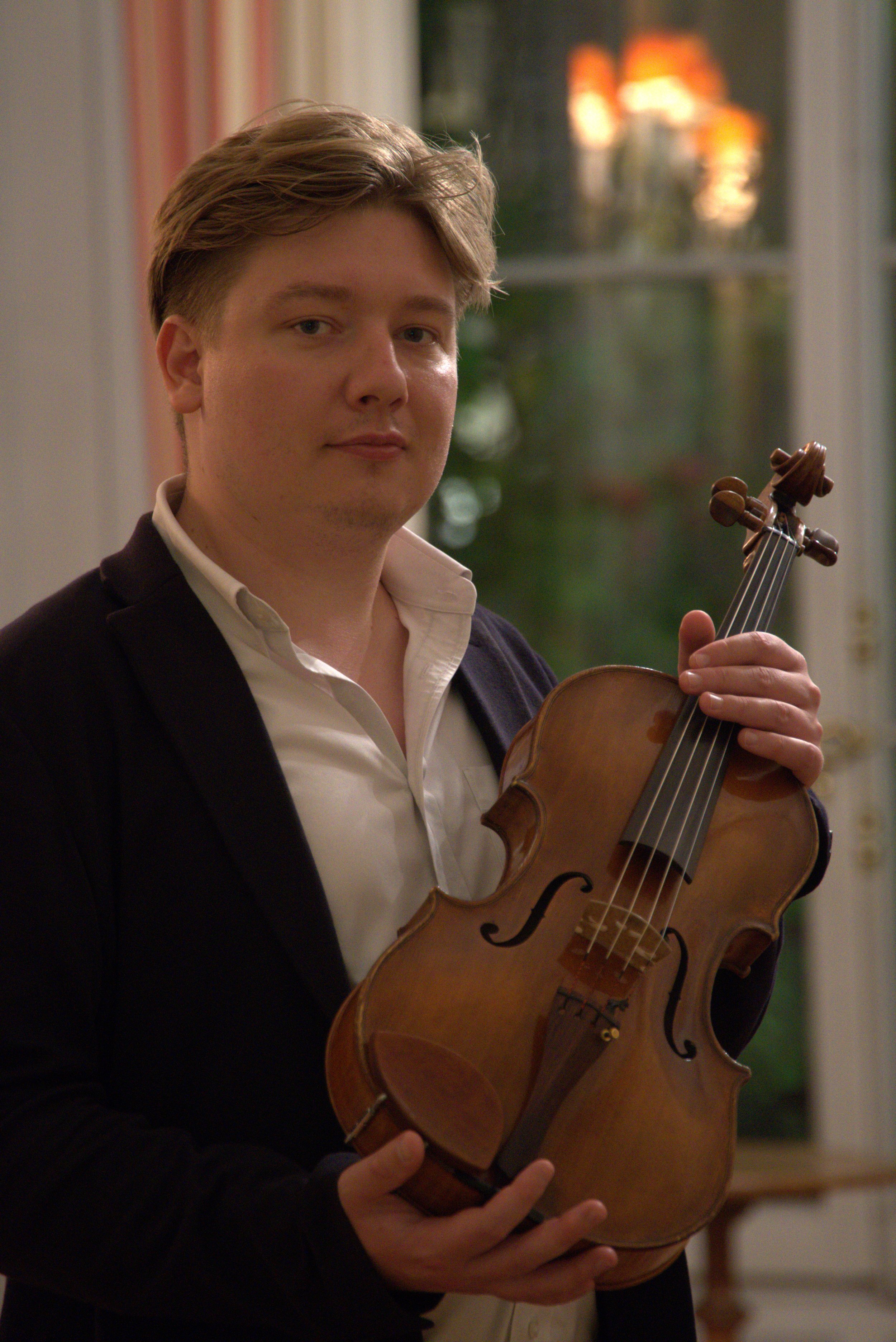 Young man in formal attire holding a violin inside a room with a window in the background.