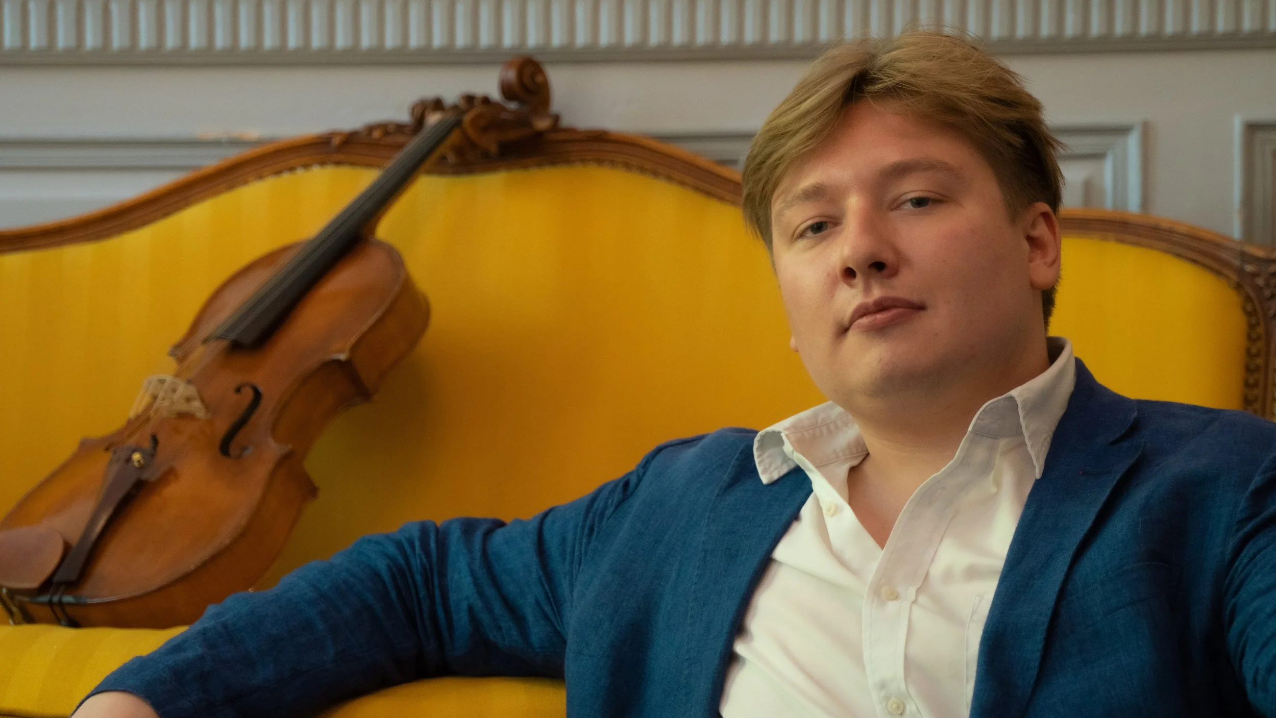 A young man with light brown hair sitting on a yellow vintage sofa with a wooden frame, next to a violin resting against the sofa.