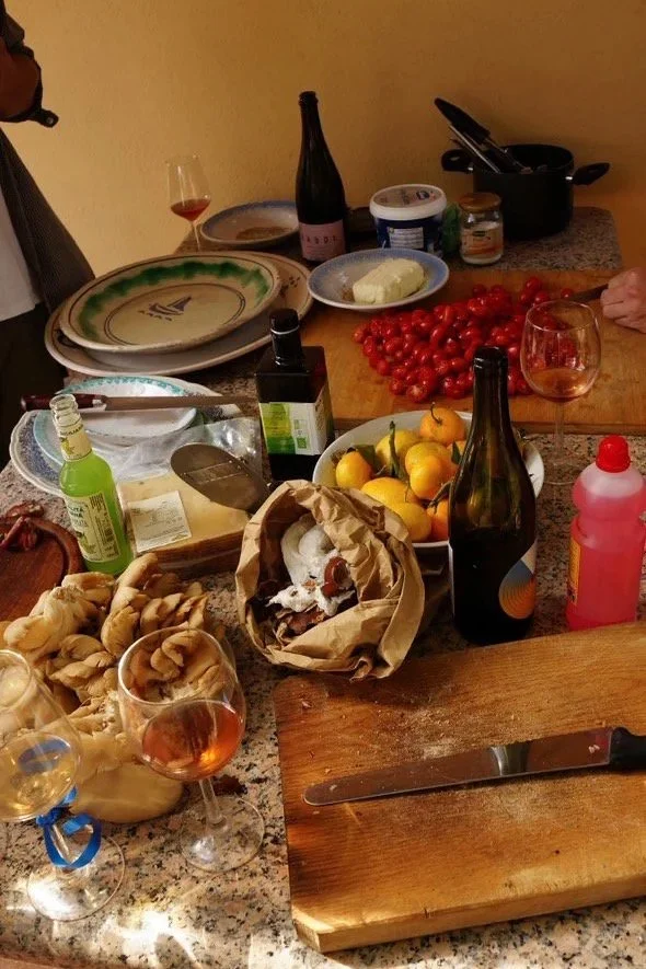 A kitchen counter with a cutting board, a large knife, and various food items including a bowl of yellow fruits, cherry tomatoes, and a paper bag with bread. There are also several glasses of rosé wine, a bottle of wine, a bottle of soda, a bottle of