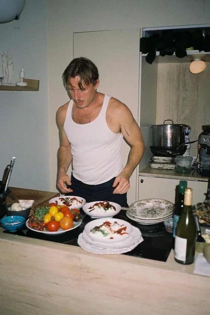 A man in a white tank top standing in a kitchen, preparing or presenting food on plates, with various fruits and kitchen items on the counter.