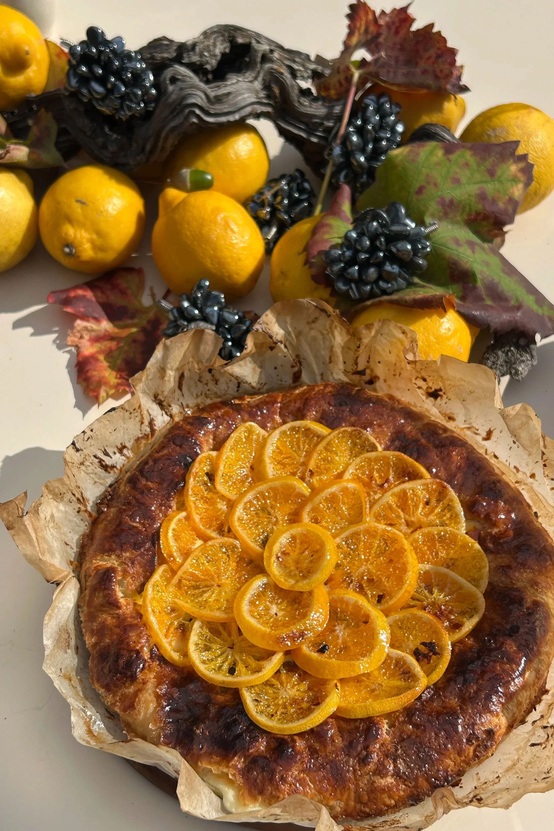 Lemon tart topped with sliced candied lemons, surrounded by lemons, black grapes, and grapevine leaves with berries and artisanal wood piece in the background.