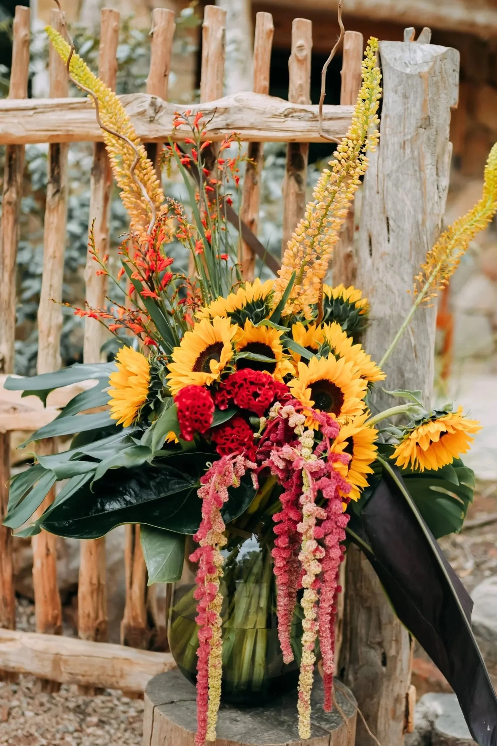 A vibrant bouquet of sunflowers, red carnations, pink and yellow amaranth, and other mixed flowers in a glass vase placed on a wooden stand outdoors.