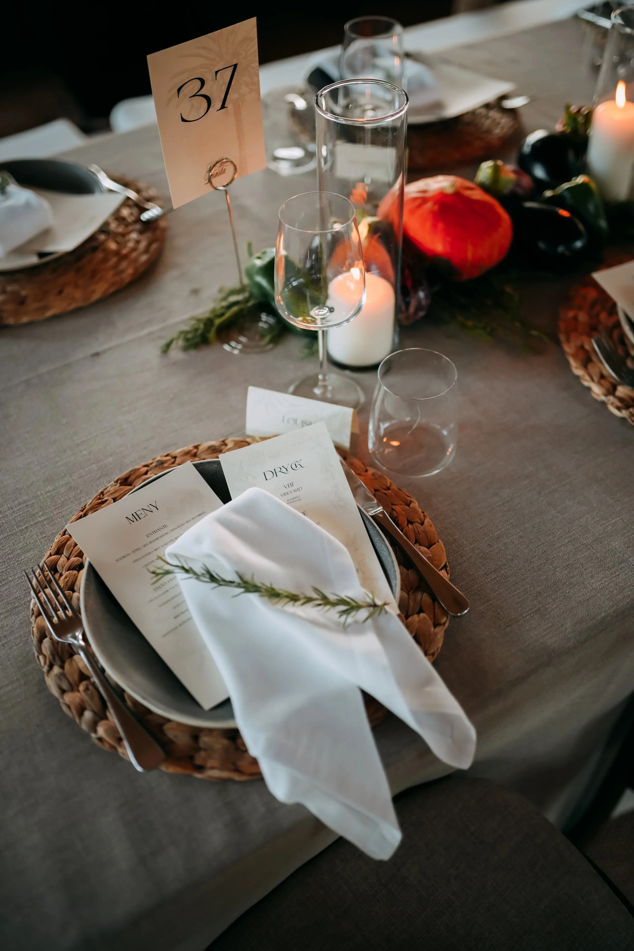 Elegant table setting with a woven placemat, white napkin with a sprig of rosemary, printed menus, wine glass, water glass, a candle, and a table number card reading '37' for a formal event or dinner.
