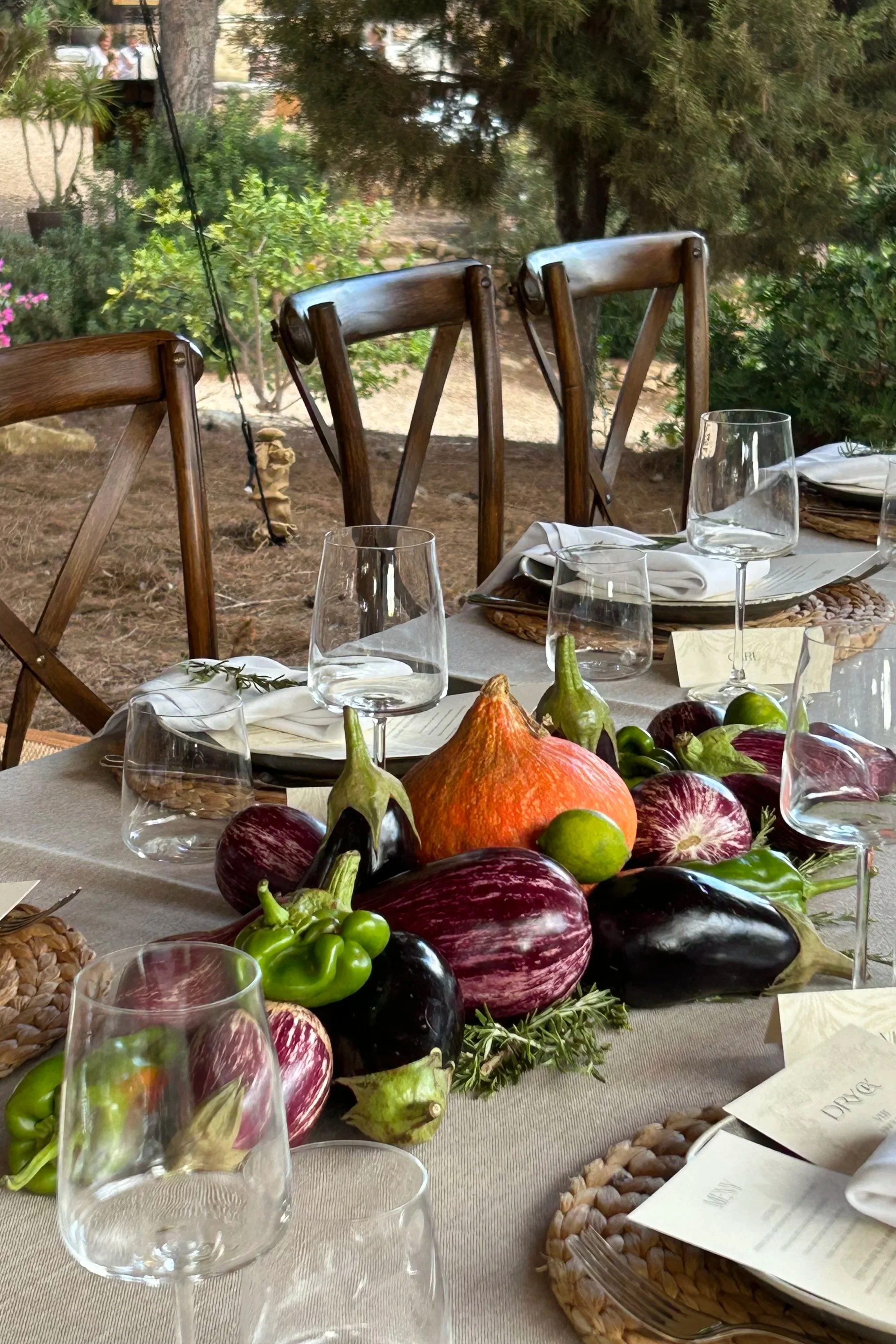 A table set outdoors with a centerpiece of eggplants, peppers, a pumpkin, limes, and greenery, surrounded by empty wine glasses, plates, napkins, and menus.