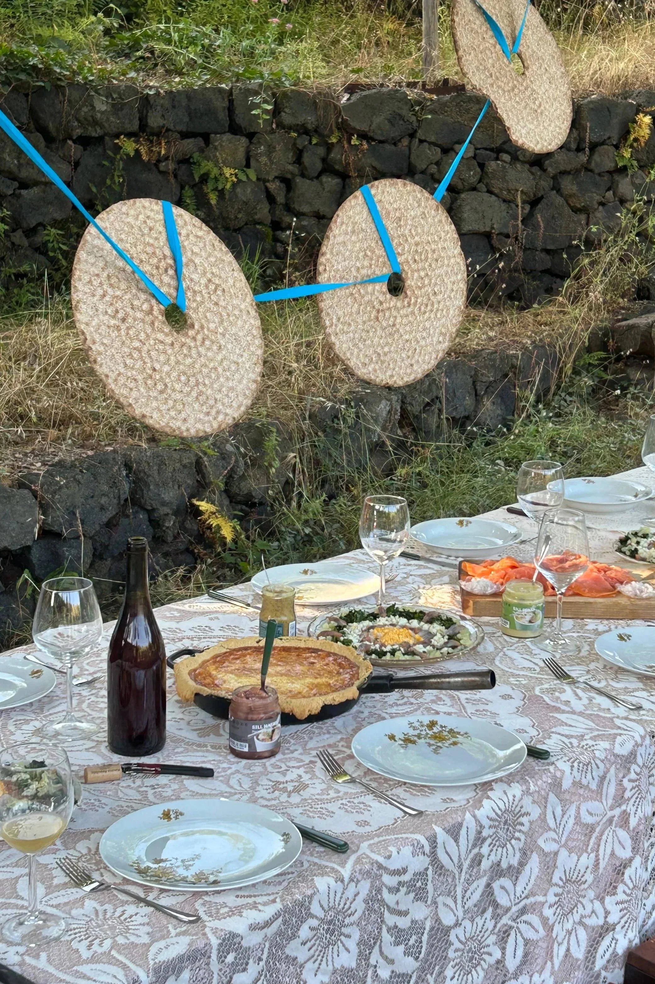 An outdoor dining table set with plates, glasses, cutlery, and various foods, against a stone wall decorated with large round bread hanging with blue ribbons.