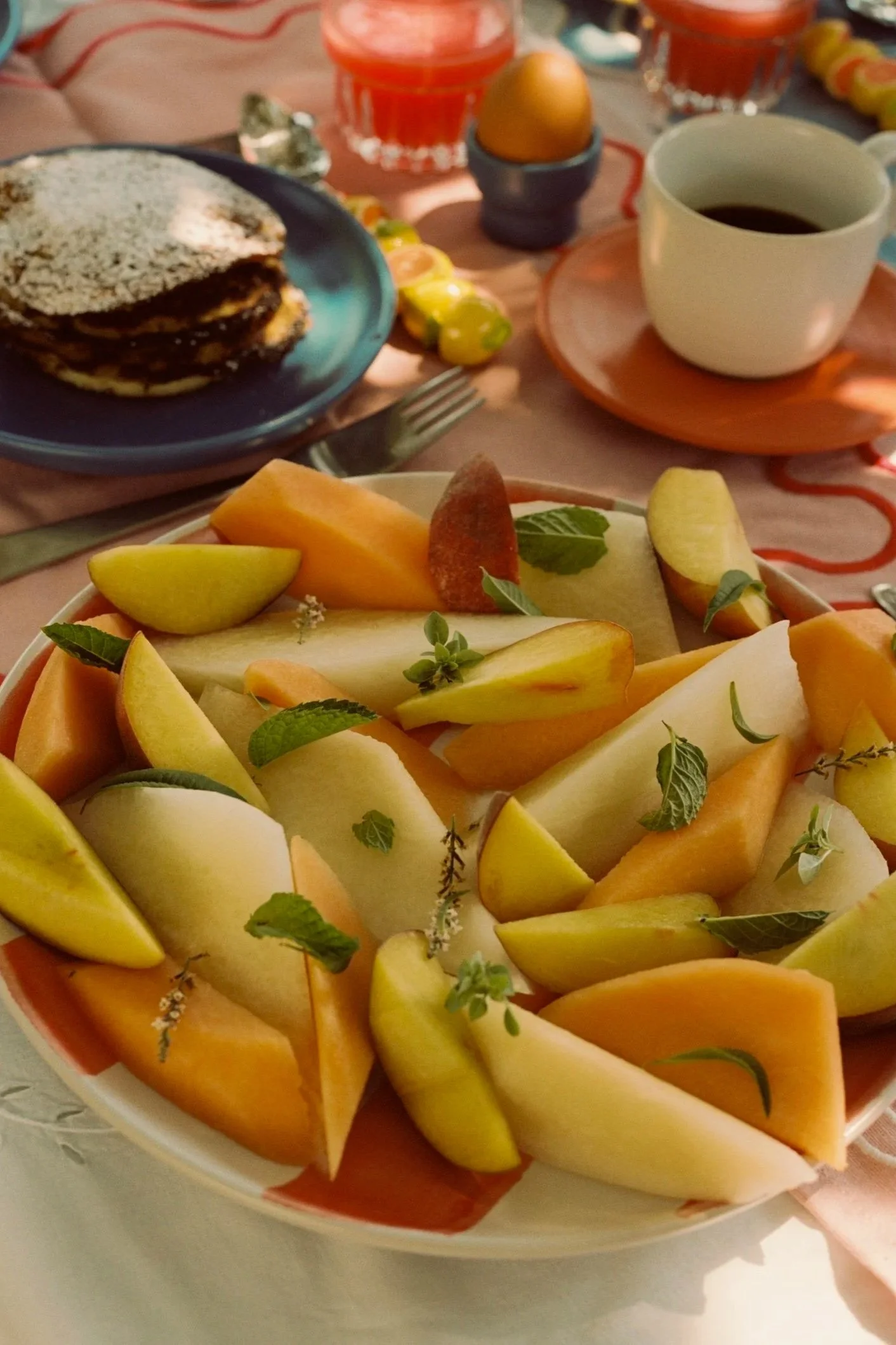 A bowl of assorted sliced melons garnished with fresh mint leaves on a table set for breakfast.