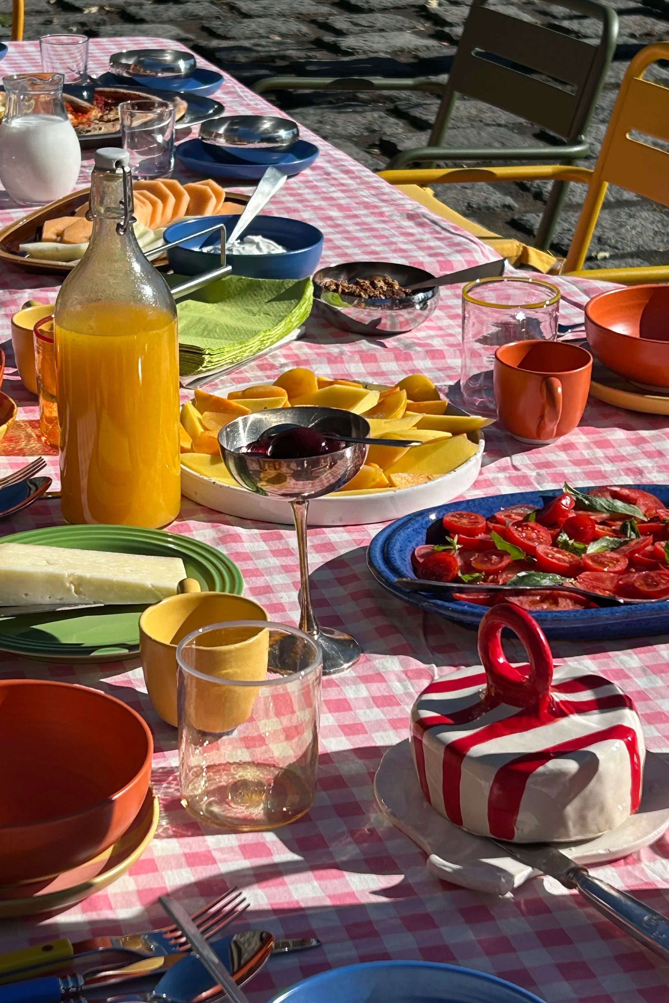 A table set for a meal outdoors with various dishes including sliced tomatoes, cheeses, fruits, drinks, and a striped ceramic cake with a loop handle on a pink checkered tablecloth.