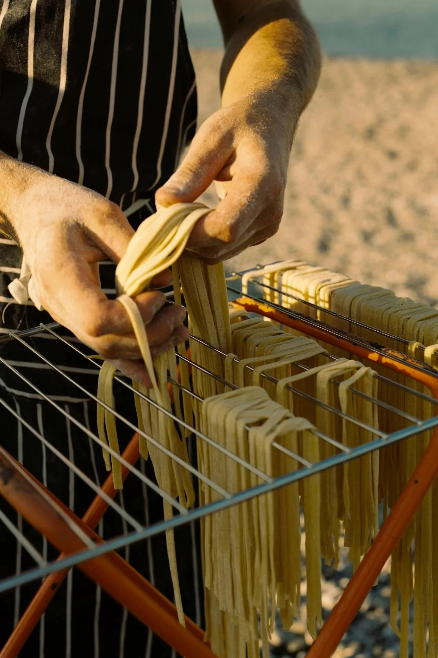 Person hanging freshly made pasta noodles on a rack outdoors.