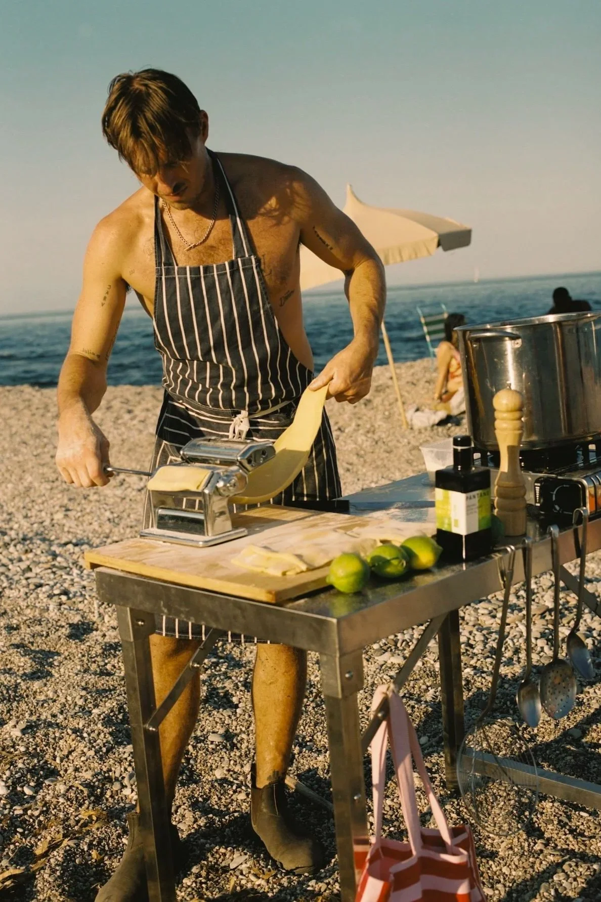 A shirtless man wearing a striped apron and black boots is preparing food on a metal table at the beach. The table holds green lemons, a pasta machine, and cooking ingredients. There is a large pot on a portable stove. The beach has pebbles, umbrella