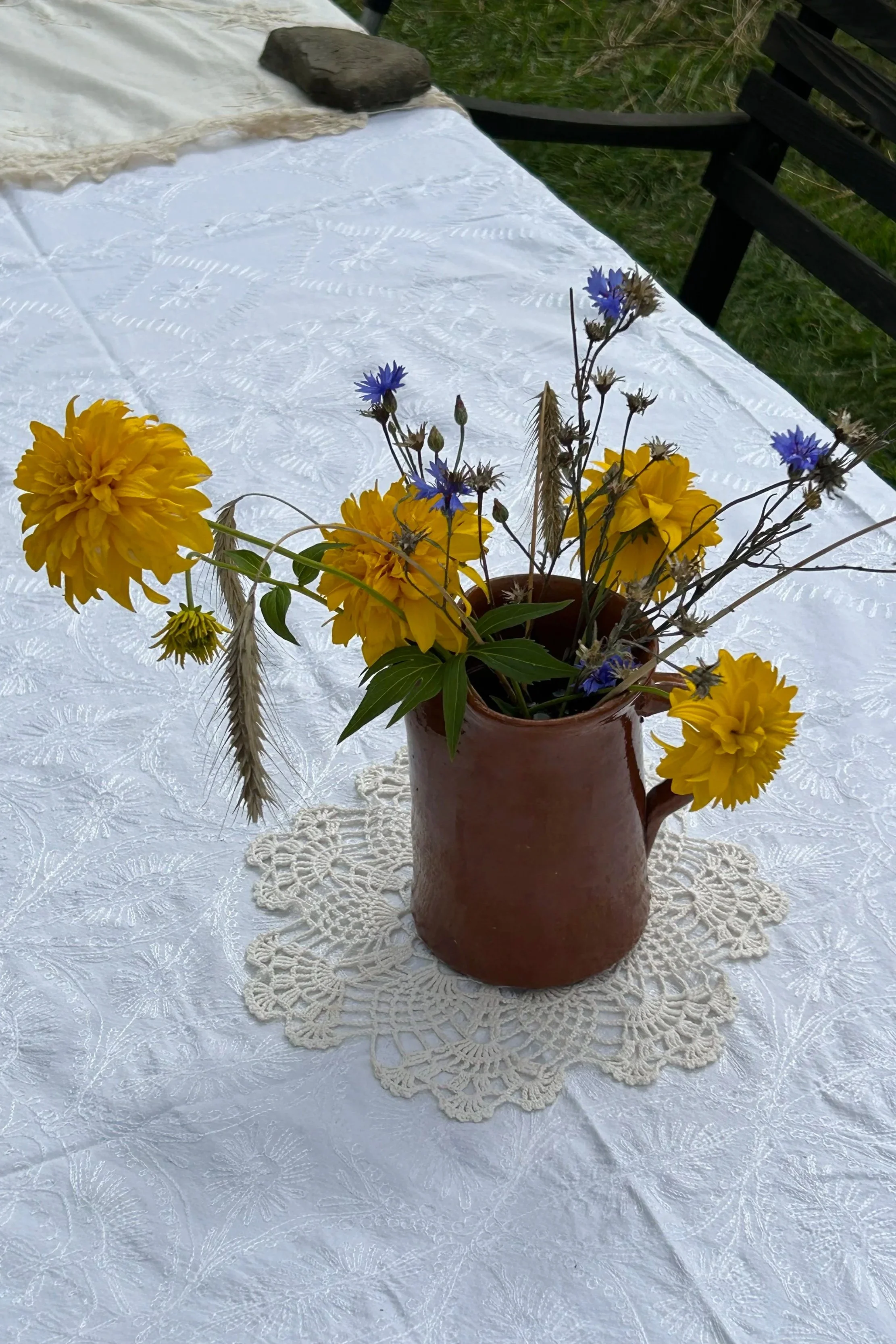A brown ceramic pitcher filled with yellow and blue flowers on a white lace tablecloth outdoors.