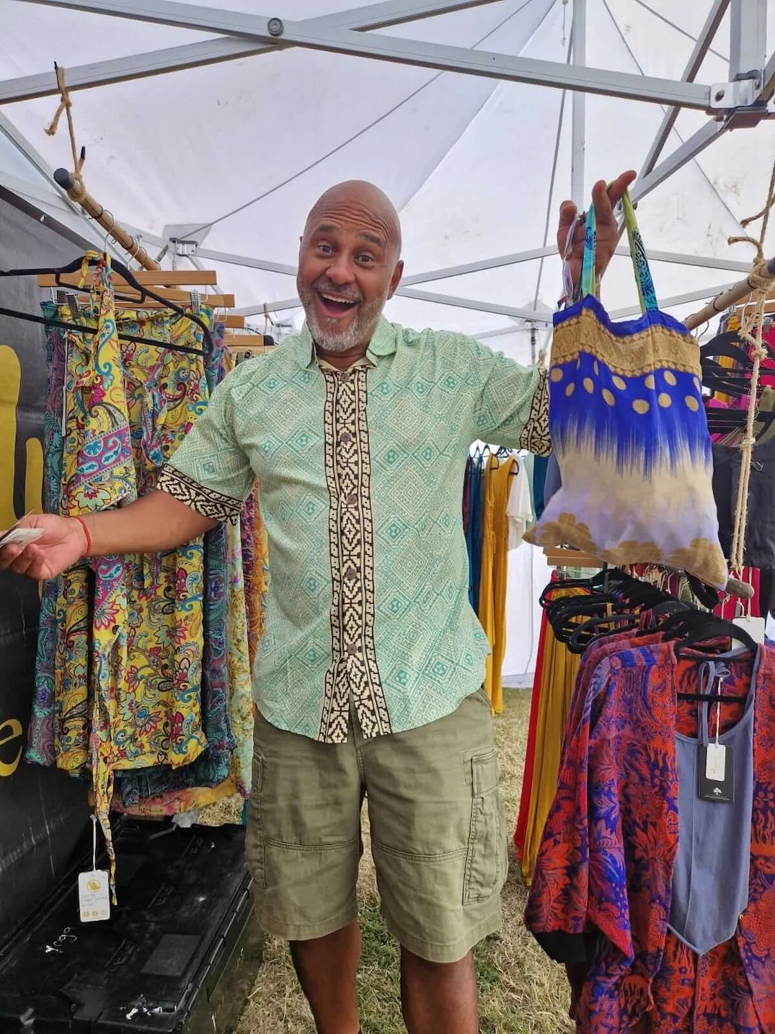 A middle-aged man smiling and holding a colorful bag inside a clothing stall with colorful garments hanging around, under a white tent roof.