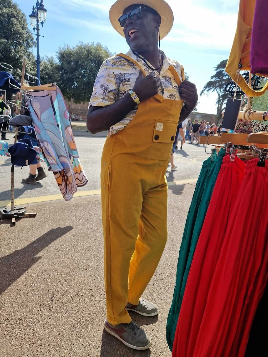 A man wearing a straw hat, sunglasses, a patterned shirt, and yellow overalls is standing outdoors at a market, smiling and holding onto his straps. There are market stalls with colorful clothing and a street scene in the background.